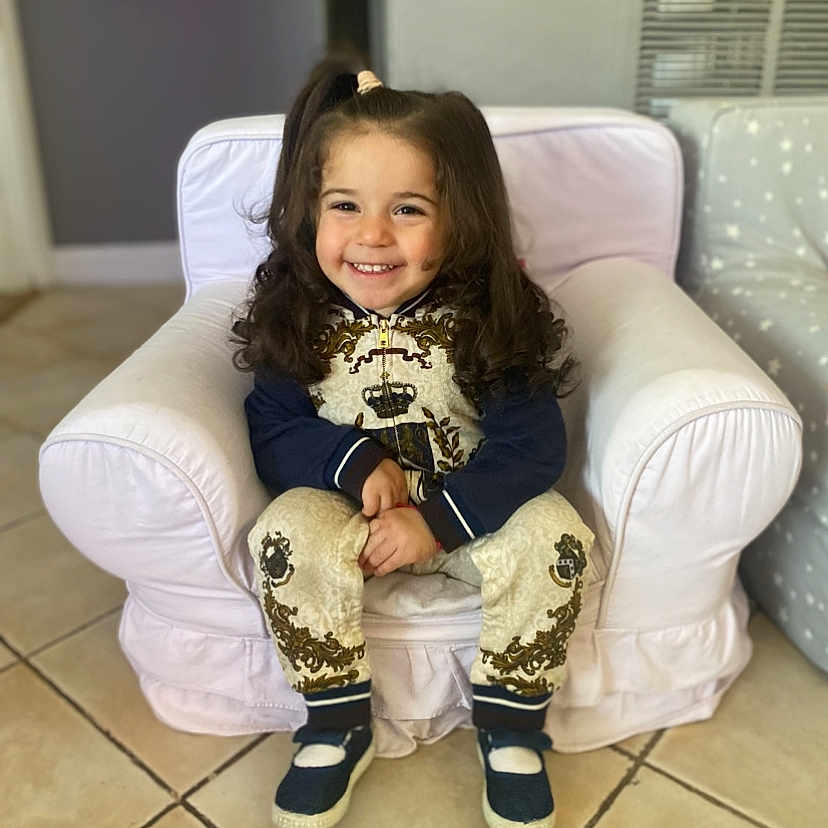 Jolie Elias is registered to the contest to win money with this photo: armchair, casual_clothing, child, cozy, curly_hair, cute, decor, dimples, floor_tiles, footwear, furniture, hair_tie, happy, indoor, person, portrait, sitting, smiling, toddler, young