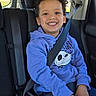 child, smiling, car_seat, seatbelt, curly_hair, hoodie, purple_hoodie, skull_design, car_interior, window, sunlight, happy, sitting, young, boy, casual_clothing, safety, daytime, portrait, vehicle