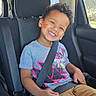 child, smiling, car_seat, seatbelt, vehicle, window, casual_clothing, face_sticker, happy, curly_hair, boy, indoor, sunlight, safety, car_interior, seat, transportation, portrait, young, kid