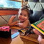 toddler, child, smile, dimples, pony_tail, table, wooden_table, restaurant, menu, keys, sweetener_packets, window, sunlight, chair, happy, indoor, person, face, arm, casual