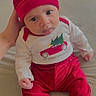 baby, infant, child, red_clothing, hat, tongue_out, cute, hand, sitting, indoors, soft_surface, playful, festive, long_sleeve_shirt, pants, person, portrait, young_child, cozy, expression