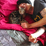 Mema is registered to the contest to win money with this photo: person, cat, bed, red_blanket, gray_bedspread, tattoo, hat, indoor, resting, snuggling, tabby_cat, human, sleeping, comfort, relaxation, pet, companion, casual_clothing, hand, face