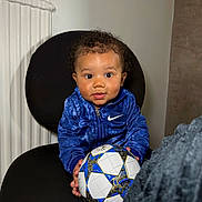 Raïley a rejoint le concours — aidez-le/la à gagner de superbes lots ! toddler, child, blue_jacket, nike, soccer_ball, black_chair, indoor, curly_hair, looking_at_camera, holding_object, seated, person, cute, portrait, smiling, young_child, home_interior, radiator, clothing, playful