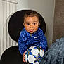 toddler, child, blue_jacket, nike, soccer_ball, black_chair, indoor, curly_hair, looking_at_camera, holding_object, seated, person, cute, portrait, smiling, young_child, home_interior, radiator, clothing, playful