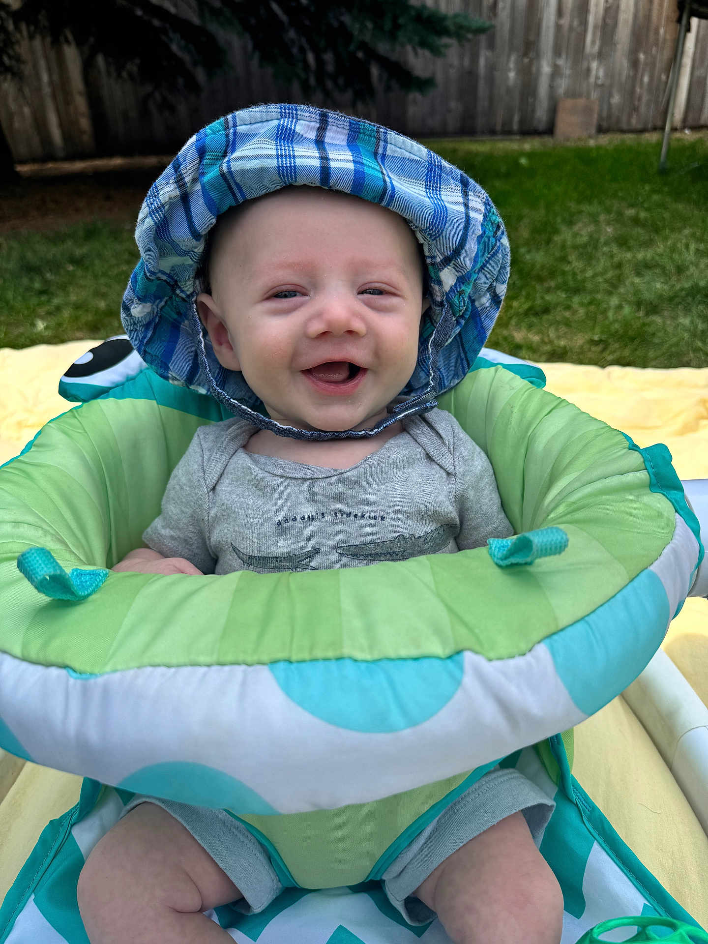Rowan joined the competition — help win amazing prizes! baby, smiling, hat, blue_hat, plaid, baby_seat, green, blue, outdoor, grass, fence, blanket, yellow_blanket, happy, infant, cute, summer, sun_hat, child, person