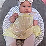 baby, infant, child, bow_headband, yellow_dress, white_tights, chevron_seat, baby_seat, pink_wall, gray_cushion, portrait, indoors, cute, chubby_cheeks, sitting, hands, feet, clothing, adorable, closeup