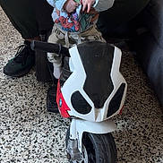 Giulianno a rejoint le concours — aidez-le/la à gagner de superbes lots ! baby, child, toy_motorcycle, indoor, floor, person, hands, superhero_clothing, sitting, supporting_hand, footwear, couch, speckled_floor, wide_eyes, curious, motorcycle_toy, white_clothing, adult, playtime, indoor_lighting
