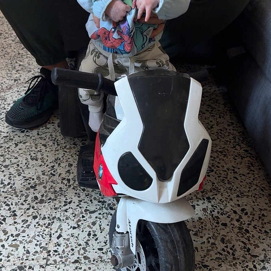 Giulianno a rejoint le concours — aidez-le/la à gagner de superbes lots ! adult, baby, child, couch, curious, floor, footwear, hands, indoor, indoor_lighting, motorcycle_toy, person, playtime, sitting, speckled_floor, superhero_clothing, supporting_hand, toy_motorcycle, white_clothing, wide_eyes