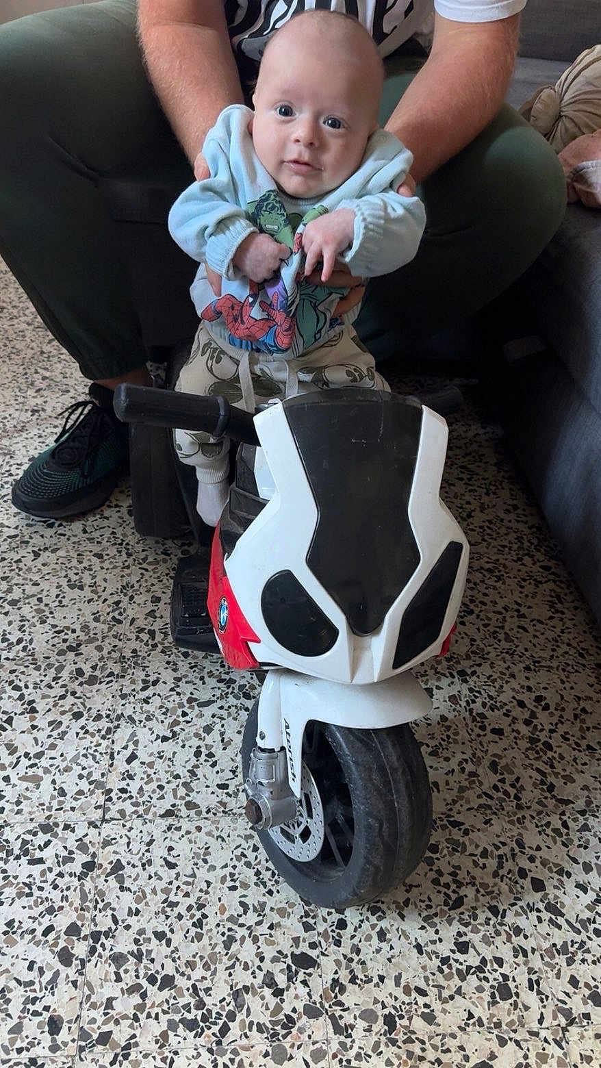 Giulianno a rejoint le concours — aidez-le/la à gagner de superbes lots ! baby, child, toy_motorcycle, indoor, floor, person, hands, superhero_clothing, sitting, supporting_hand, footwear, couch, speckled_floor, wide_eyes, curious, motorcycle_toy, white_clothing, adult, playtime, indoor_lighting