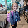 child, smiling, indoor, backpack, girl, casual_clothing, shoes, tiled_floor, furniture, couch, shelves, decor, buddha_art, window, door, household_items, person, young_child, happy, standing