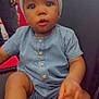 baby, child, denim_romper, headwrap, chair, indoor, curious, sitting, cute, portrait, person, black_chair, buttons, footwear, skin, hand, face, eyes, expression, young_child