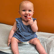 Harley is registered to the contest to win money with this photo: baby, child, pacifier, blue_dress, sitting, medical_table, orange_wall, smiling, happy, infant, cute, cheerful, barefoot, skin, indoor, young_child, portrait, baby_girl, clothing, hand