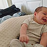 baby, infant, smile, tongue_out, pillow, blanket, clothing, onesie, indoor, door, bag, happy, cute, person, child, lying_down, soft_texture, home, casual, relaxed