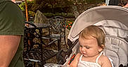 Isabella is registered to the contest to win money with this photo: baby, casual_clothing, child, curious, daylight, garden, greenery, metal_chair, outdoor, park, person, plastic_wrap, relaxing, shoes, striped_umbrella, stroller, summer, table, toddler, umbrella