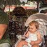 Isabella is registered to the contest to win money with this photo: toddler, stroller, child, outdoor, umbrella, striped_umbrella, plastic_wrap, greenery, metal_chair, table, person, casual_clothing, shoes, baby, summer, daylight, park, curious, relaxing, garden
