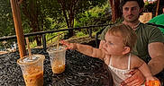 Isabella joined the competition — help win amazing prizes! child, toddler, man, outdoor, table, iced_coffee, drink, umbrella, greenery, tree, fringe, summer, sunlight, curious, reaching, holding, plastic_cup, beverage, casual_clothing, relaxing