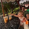 child, toddler, man, outdoor, table, iced_coffee, drink, umbrella, greenery, tree, fringe, summer, sunlight, curious, reaching, holding, plastic_cup, beverage, casual_clothing, relaxing