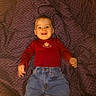 Léon a rejoint le concours — aidez-le/la à gagner de superbes lots ! baby, child, infant, smiling, red_shirt, blue_jeans, blanket, patterned_blanket, lying_down, happy, tongue_out, indoor, portrait, cute, clothing, person, face, young_child, casual_wear, warm_lighting