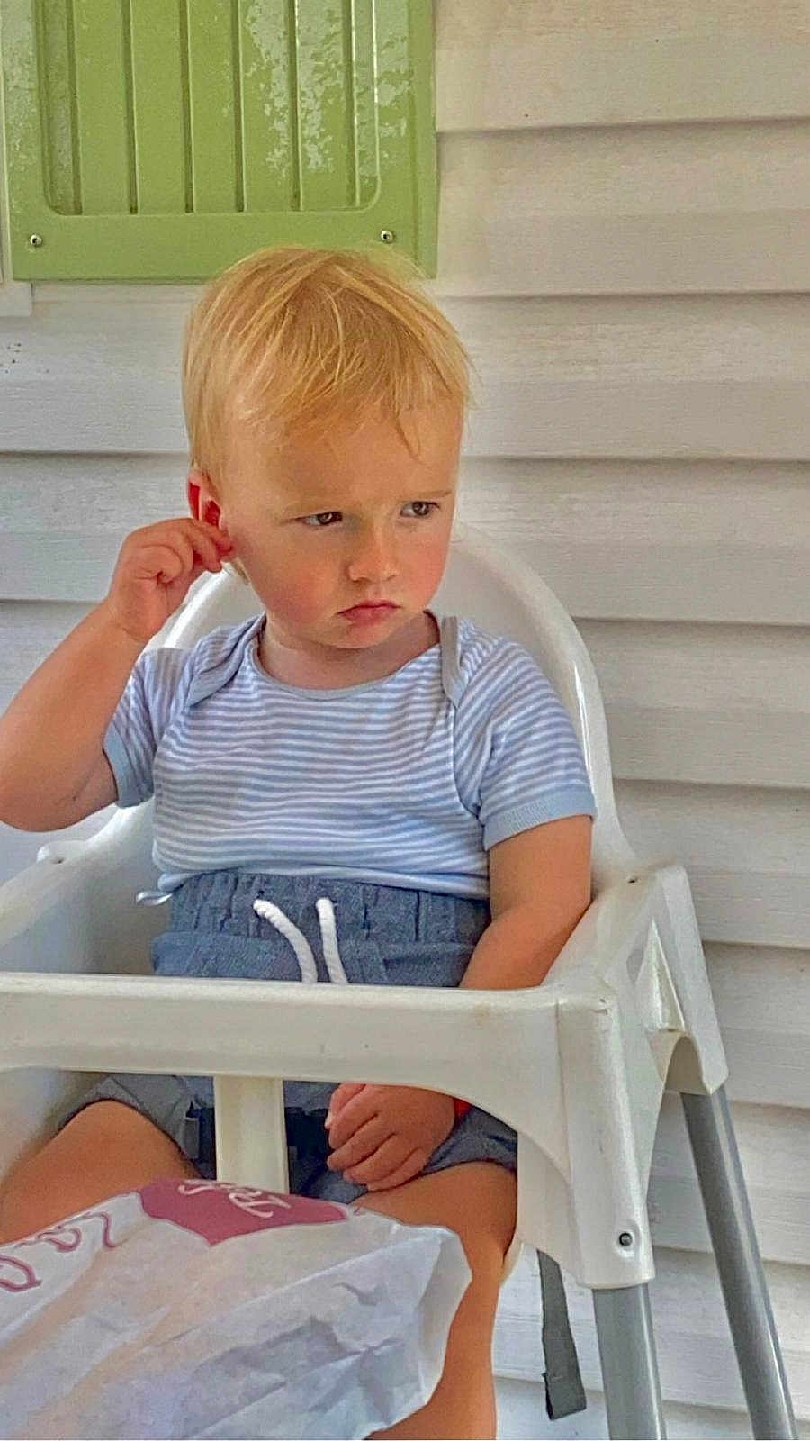 Alan a rejoint le concours — aidez-le/la à gagner de superbes lots ! toddler, child, high_chair, striped_shirt, blue_shorts, serious_expression, scratching_ear, white_wall, green_vent, paper_bag, indoor, furniture, person, seated, short_hair, casual_clothing, young_child, contemplative, blond_hair, portrait