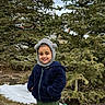 child, smiling, outdoor, winter_clothing, hat, jacket, trees, evergreen, snow, grass, nature, person, young, cute, cold_weather, green_pants, standing, happy, face, portrait