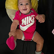 Leya a rejoint le concours — aidez-le/la à gagner de superbes lots ! baby, smiling, onesie, nike, red_clothing, sock, footwear, cushion, yellow, black_background, infant, indoor, happy, cute, portrait, child, person, seated, looking_at_camera, clothing