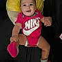 baby, smiling, onesie, nike, red_clothing, sock, footwear, cushion, yellow, black_background, infant, indoor, happy, cute, portrait, child, person, seated, looking_at_camera, clothing