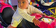 Leya participe au concours pour gagner de l'argent avec cette photo : baby, child, arcade, motorcycle, pink, headband, bow, white_clothing, indoor, game_machine, neon_lights, colorful, toy, playing, person, footwear, curious, seat, hand, looking