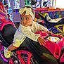 baby, child, arcade, motorcycle, pink, headband, bow, white_clothing, indoor, game_machine, neon_lights, colorful, toy, playing, person, footwear, curious, seat, hand, looking