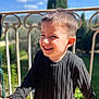 child, boy, smiling, outdoor, sunlight, sweater, black_clothing, happy, portrait, face, short_hair, railing, nature, greenery, daylight, young, person, casual_wear, background_blur, cheerful
