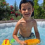 child, boy, pool, water, water_toys, inflatable_pool, summer, outdoor, sunny, blue_sky, plants, greenery, cap, swimwear, smiling, playing, bubbles, yellow, orange, leisure