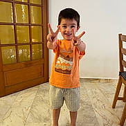 Naël a rejoint le concours — aidez-le/la à gagner de superbes lots ! child, boy, smiling, peace_sign, barefoot, orange_shirt, shorts, indoor, marble_floor, wooden_door, chair, happy, standing, casual_clothing, home, person, portrait, cute, young, playful