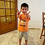 child, boy, smiling, peace_sign, barefoot, orange_shirt, shorts, indoor, marble_floor, wooden_door, chair, happy, standing, casual_clothing, home, person, portrait, cute, young, playful