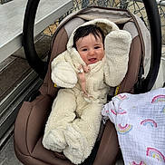 Emerson joined the competition — help win amazing prizes! baby, child, car_seat, fluffy_onesie, mittens, smiling, happy, blanket, rainbow_pattern, chain_link_fence, metal_bleachers, outdoor, infant, cute, seated, pacifier, black_hair, warm_clothing, playful, portrait