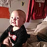 Drakken is registered to the contest to win money with this photo: baby, child, infant, smiling, happy, portrait, sitting, bed, bedroom, indoors, clothing, black_shirt, face, eyes, head, skin, blanket, pillow, wall, laundry