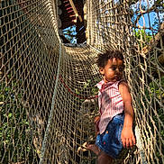 Leya a rejoint le concours — aidez-le/la à gagner de superbes lots ! toddler, child, rope_bridge, playground, outdoor, forest, sunlight, netting, barefoot, denim_skirt, pink_shirt, curly_hair, tree, nature, daylight, play, adventure, young_child, summer_clothing, exploration