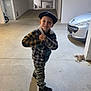 beret, black_shoes, boy, camo_pants, car, casual_clothing, child, concrete_floor, concrete_wall, hands_clasped, indoor, lighting, parking_garage, plaid_jacket, pose, shadow, smile, standing, vehicle, young_person