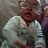 Josiah joined the competition — help win amazing prizes! baby, child, infant, glasses, oversized_glasses, face, portrait, sitting, couch, indoor, home, clothing, onesie, zipper, sleeves, close_up, funny, looking_at_camera, head, photograph