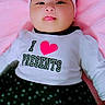 baby, infant, headband, bow, tongue, shirt, text, polka_dot, skirt, pink_blanket, lying_down, cute, child, adorable, clothing, portrait, face, person, indoors, smiling