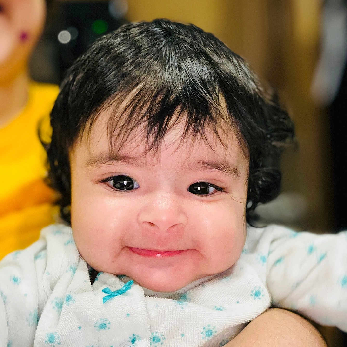 Maia is registered to the contest to win money with this photo: baby, black_hair, blue_dots, child, close_up, clothing, curly_hair, cute, face, happy, holding, indoor, infant, person, portrait, skin, smile, warm_light, white_outfit, young_child