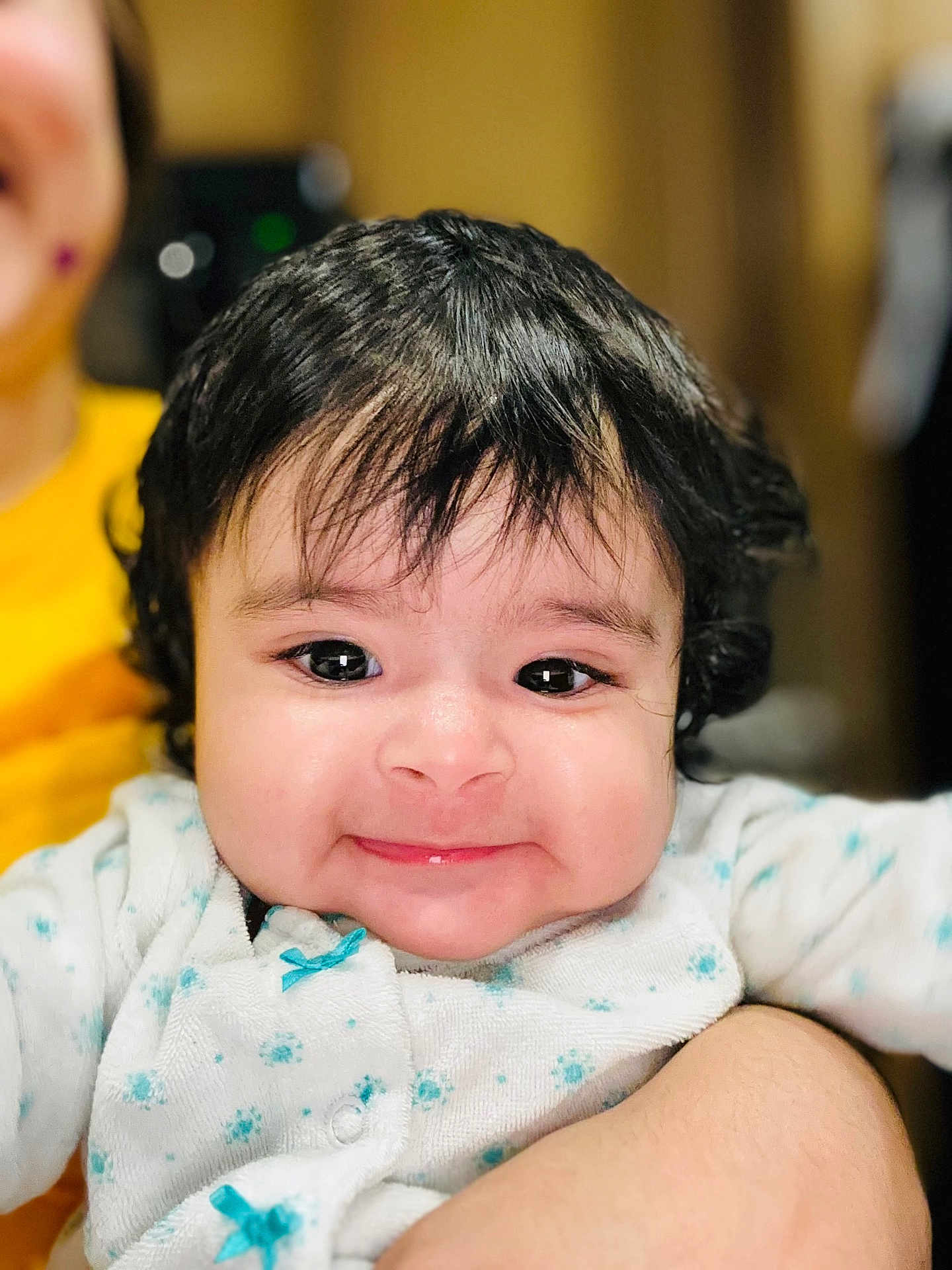 Maia is registered to the contest to win money with this photo: baby, child, face, smile, black_hair, curly_hair, infant, cute, portrait, clothing, white_outfit, blue_dots, close_up, person, holding, skin, indoor, happy, young_child, warm_light