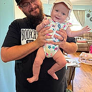 Ivy is registered to the contest to win money with this photo: baby, man, glasses, hat, pikachu, onesie, smile, beard, living_room, furniture, curtains, window, wood_floor, table, person, child, indoor, holding, happy, home
