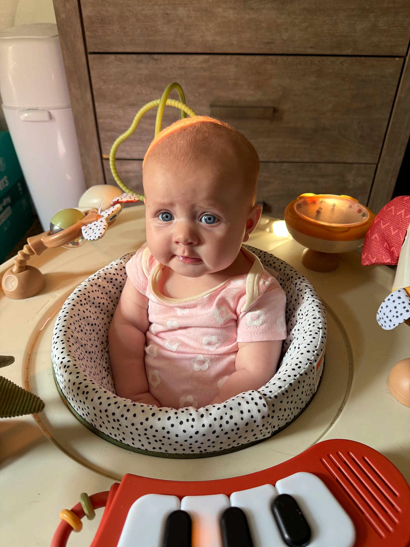 Ivy joined the competition — help win amazing prizes! baby, infant, onesie, pink_clothing, polka_dot, baby_seat, toy, keyboard_toy, blue_eyes, face, expression, indoor, wooden_drawer, child, cute, puzzled, soft_lighting, play_area, head, sitting
