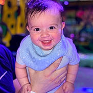Elio is registered to the contest to win money with this photo: baby, smiling, happy, child, blue_bib, onesie, hand, person, face, indoor, colorful_lights, cute, infant, portrait, holding, joy, cheerful, young_child, background_blur, human