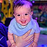 baby, smiling, happy, child, blue_bib, onesie, hand, person, face, indoor, colorful_lights, cute, infant, portrait, holding, joy, cheerful, young_child, background_blur, human