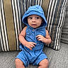 baby, child, blue_clothing, hood, whale_print, couch, striped_pillows, gray_fabric, sitting, curious_expression, indoors, infant, cute, casual_wear, portrait, small_hands, bare_legs, comfortable, soft_lighting, relaxed
