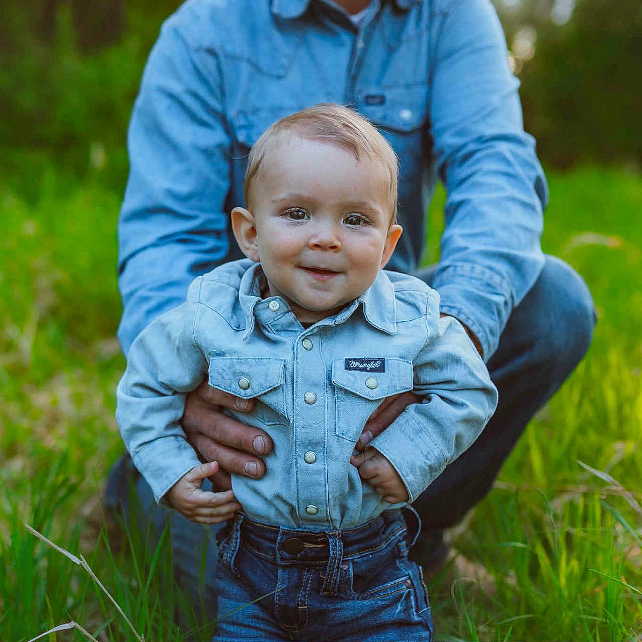 Wrye is registered to the contest to win money with this photo: adult_hands, baby, casual_clothing, child, cute, daylight, denim_shirt, field, grass, greenery, happy, jeans, nature, outdoor, person, portrait, smiling, supporting_hands, toddler, young_child