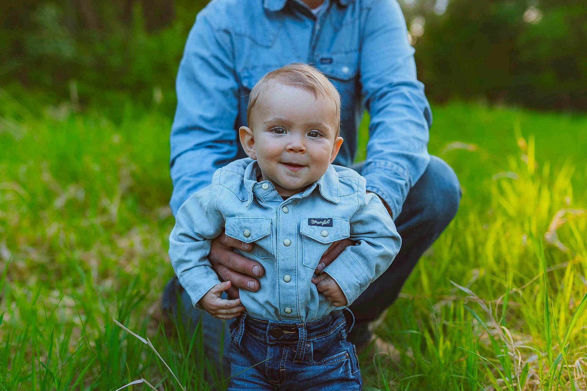 Wrye is registered to the contest to win money with this photo: toddler, child, denim_shirt, jeans, smiling, outdoor, grass, greenery, adult_hands, nature, portrait, cute, baby, person, young_child, casual_clothing, daylight, happy, supporting_hands, field
