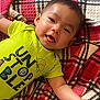 baby, child, infant, smiling, tongue_out, lying_down, plaid_blanket, blanket, green_shirt, text_on_shirt, cute, happy, portrait, face, skin, arm, indoors, cozy, toddler, clothing