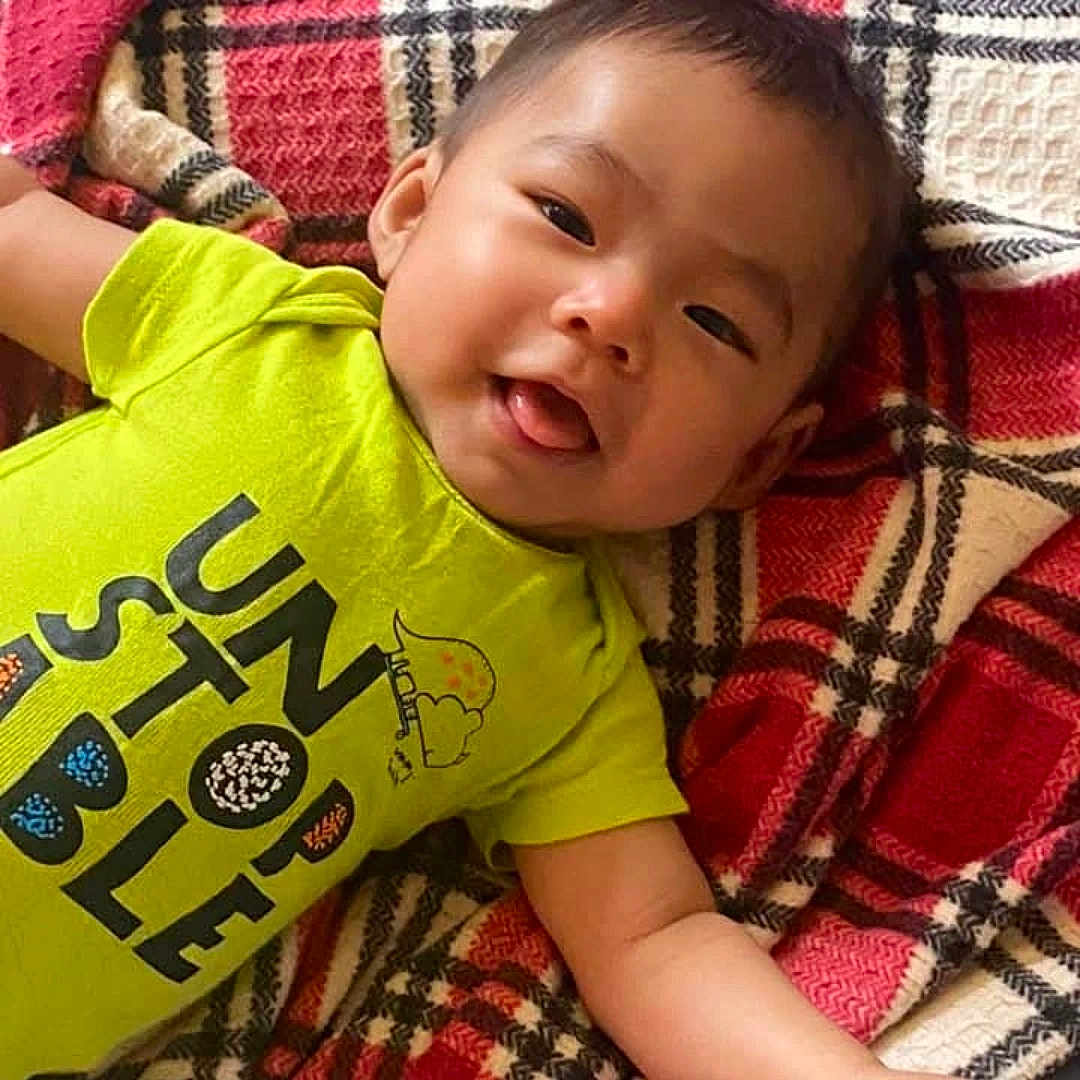 John Lee joined the competition — help win amazing prizes! baby, child, infant, smiling, tongue_out, lying_down, plaid_blanket, blanket, green_shirt, text_on_shirt, cute, happy, portrait, face, skin, arm, indoors, cozy, toddler, clothing