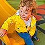 toddler, child, yellow_jacket, curly_hair, playground, slide, smile, outdoor, casual_clothing, blue_pants, play_area, happy, young_child, sitting, daylight, play_equipment, footwear, fun, park, cute